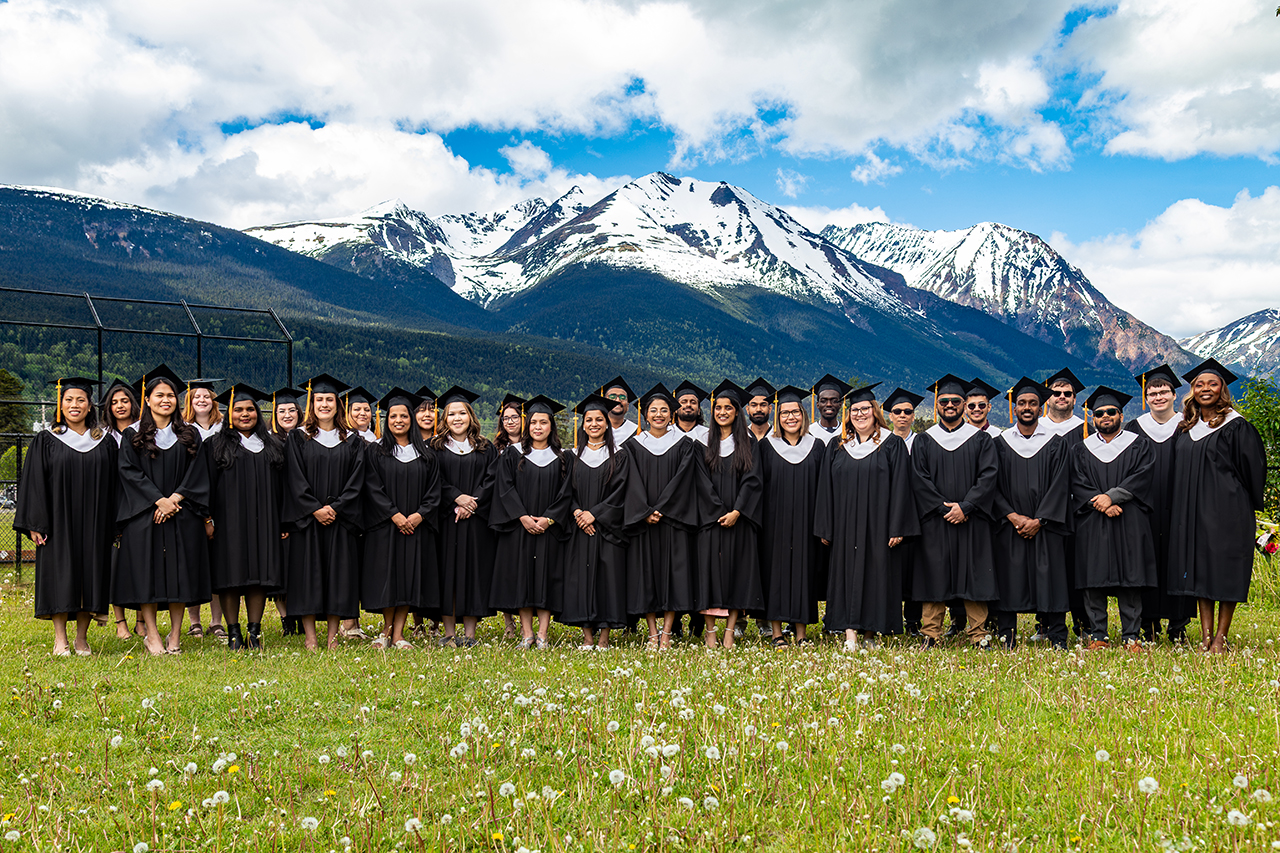 The CMTN Smithers class of 2026 poses on a lawn in their graduation caps and gowns. Dzilh Yez Mountain is in the background.